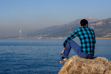 Türkiye 22 Ocak 2017: genç erkeklerde bir gömlek bir kafeste oturur güneşli bir öğleden sonra Iskenderun dağlardaki çerçevede bir mavi deniz kıyısına yakın bir taş üzerinde