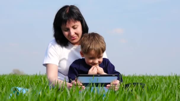 Šťastná rodina: na zelené trávě v parku odpočívejte matka a dítě. Matka a syn se dívají na obrazovku tabletu. Technologie, Internet, komunikace.