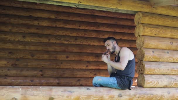 Un joven enciende un cigarro mientras está sentado en un tronco de una casa de madera . — Vídeos de Stock