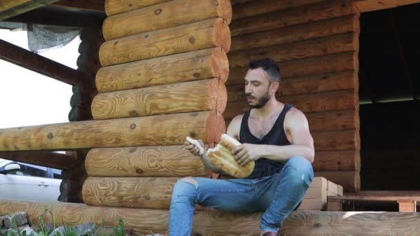 A handsome man crouched on the doorstep of the house and eats appetizing bread. — Stock Video