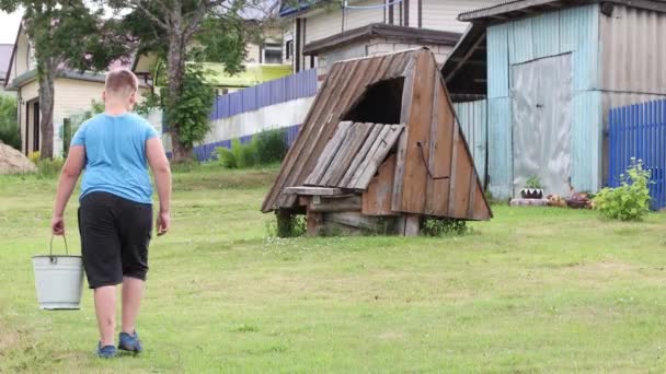 En pojke i blå tröja och shorts med en hink i handen går till brunnen. — Stockvideo