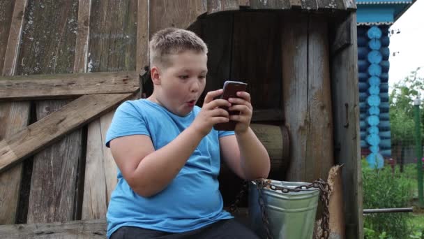 Close-up boy sitting on a wooden well plays the phone. — Stock Video