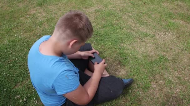 トップビューでは、緑の芝生の上に座っている男の子が感情的にスマートフォンで再生されます. — ストック動画