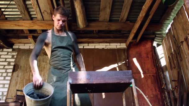 A male blacksmith in a veil pours coal from a bucket into a forge. — Stock Video