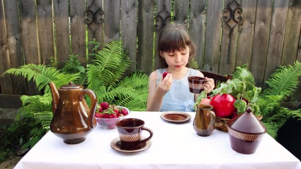 Een klein meisje drinkt thee, eet bessen, likt haar vingers. — Stockvideo