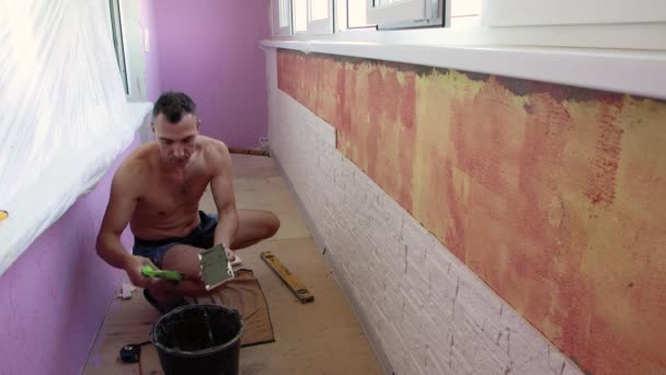 Человек на корточках кладет плитку на стену лоджии. — стоковое видео