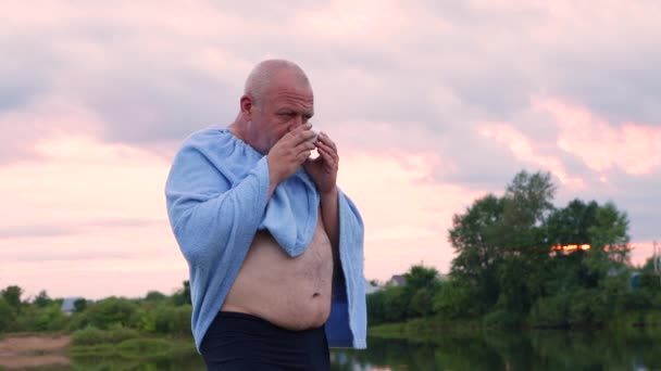 Ein Mann steht mit einem blauen Handtuch auf den Schultern am See. — Stockvideo