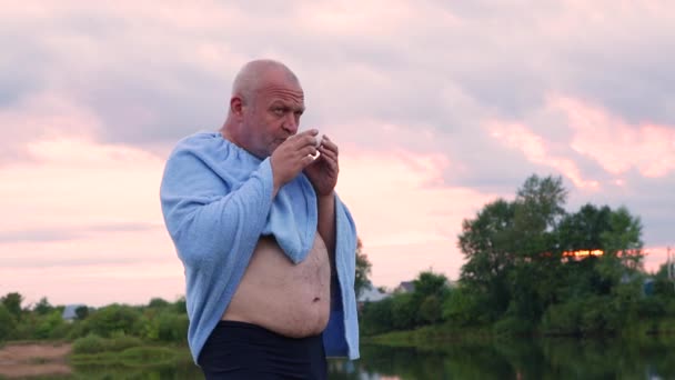 Un hombre está de pie en la orilla del lago con una copa en sus manos. — Vídeos de Stock