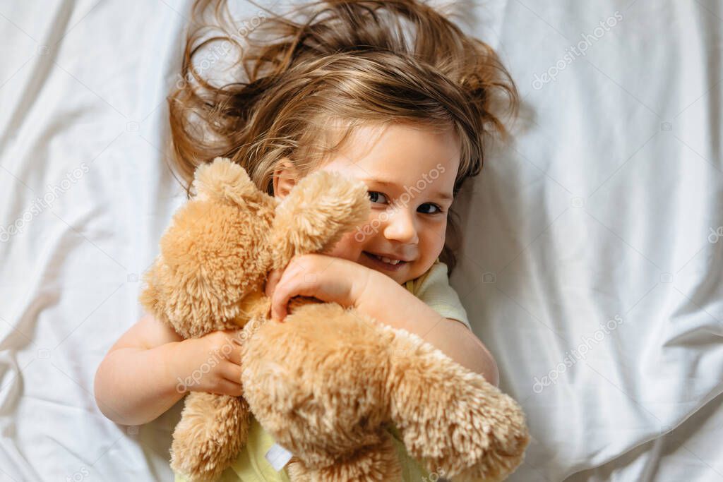 Smiling toddler girl holding teddy bear and lying in bed on white sheets at home. Happy child. Top view. 