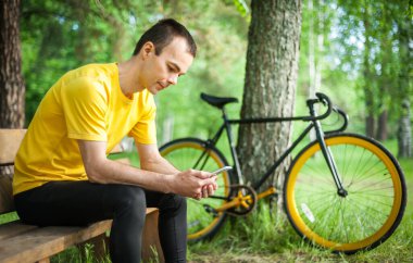 Halka açık bir parkta bankta oturan genç bir adam mobil iletişim yoluyla iletişim kuruyor. Arka planda bisikletiyle birlikte. Ağaçların ve bitkilerin arasında.