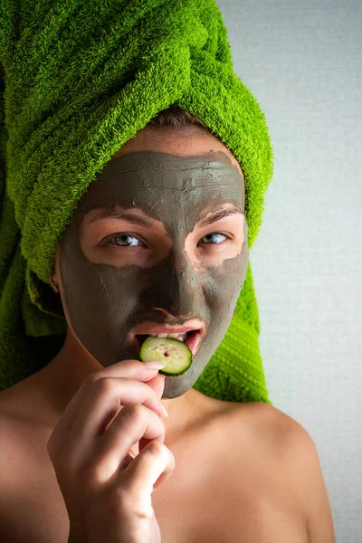 Beautiful young woman with facial mask on her face holding slices of cucumber. Skin care and treatment, spa, natural beauty and cosmetology concept.