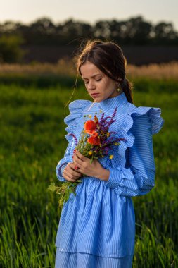 Mavi ve beyaz çizgili elbiseli Genç kadın iki elinle bir buket yaz çiçek gözleri ile kapalı ve başını bir bahar öğleden sonra boyunca güneşli hava, yeşil buğday alanında dururken ettirildiğinde tutuyor. Saçlarını flutterin