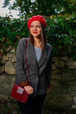 Kırmızı bere kırmızı çanta kamera yakınındaki taş çit uzak seyir ile genç bir kadın portresi bitki örtüsü ile kaplı.