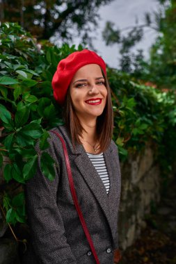 Kırmızı bere bitki örtüsü ile kaplı bir taş çit yaslanmış bir genç gülümseyen kadın portresi.