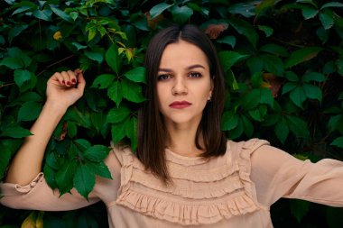 Genç kadın üzerinde yeşil yapraklarla kaplı ve kameraya bakarak bir taş çit yaslanmış.