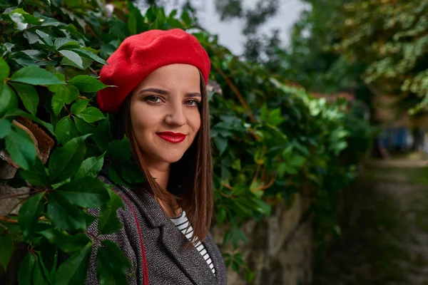 Kırmızı bere bitki örtüsü ile kaplı bir taş çit yaslanmış gülümseyen genç kadın görüntüsünü kapatmak.