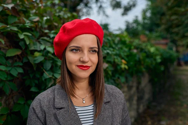 Kırmızı bere kamera bir taş çit yakınındaki uzak arıyorum Genç bir kadın portresi bitki örtüsü ile kaplı.