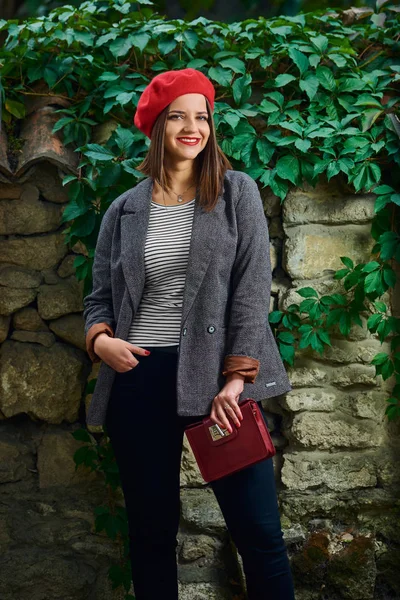 Young woman in red beret looking into camera against a stone fence with vegetation while holding a red handbag.
