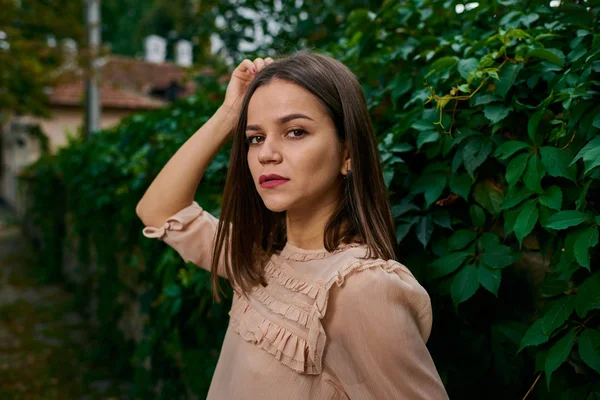 Young woman posing in a beige blouse at stone fence covered with leaves.