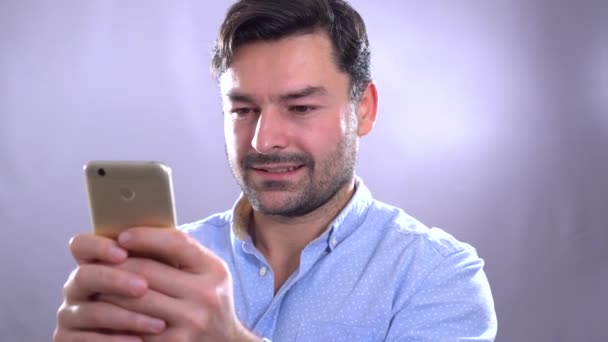 Joven sonriente sosteniendo el teléfono inteligente y mirándolo. Retrato de un hombre feliz usando teléfono móvil. Comunicación, beige . — Vídeo de stock