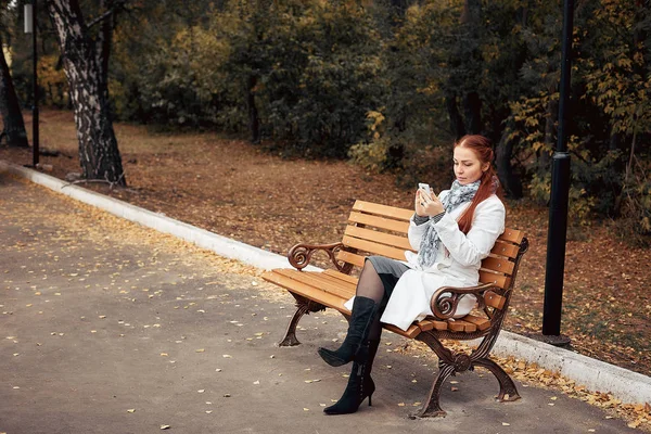 red-haired middle-aged woman with a smartphone in her hand sits on a bench in the Park and communicates in social networks.