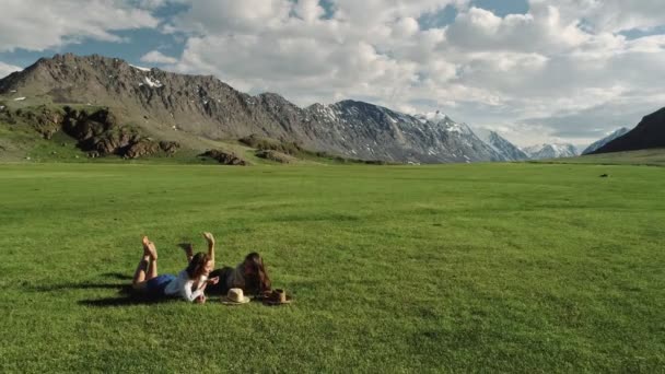 Дві щасливі друзі лежать на зеленій траві на тлі гір — стокове відео