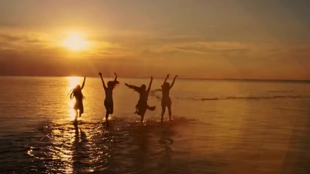 Skupina happy dívek běží a hrát ve vodě na pláži na tanec slunce, postřik nad letní západ slunce. Beach party. Sluneční záře. Zpomalený pohyb — Stock video