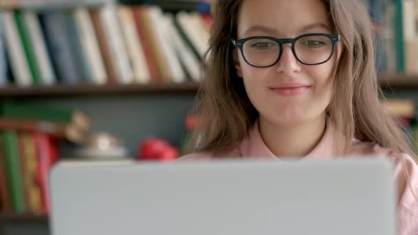 Vrouwelijke student zitten tegen boekenplank en met behulp van laptop in de bibliotheek — Stockvideo