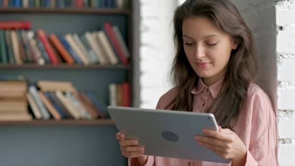 Primer plano de la joven rizada con gafas que trabajan en el dispositivo de la tableta y sonriendo a la cámara en el pasillo de la biblioteca. Retrato. En interiores — Vídeos de Stock