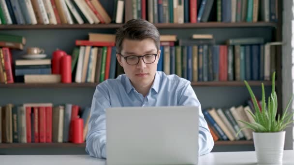 Male student sitting against bookshelf and using laptop in the library — Stock Video