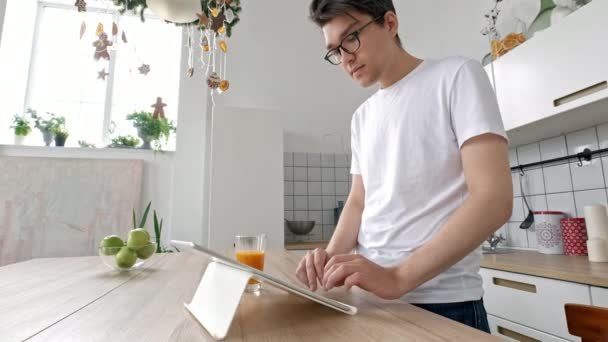 Vonzó férfi otthon segítségével tabletta konyhában küldött üzenetet a szociális média, mosolyog, élvezi a modern életmód, fehér inget visel — Stock videók