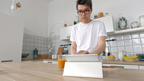 Hombre atractivo en casa usando tableta en la cocina enviando un mensaje en las redes sociales sonriendo disfrutando de un estilo de vida moderno usando camisa blanca — Vídeo de stock