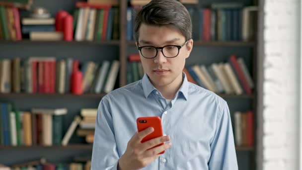 Молодой человек портрет с помощью смартфона смеется стоя в библиотеке социальные медиа смешно — стоковое видео