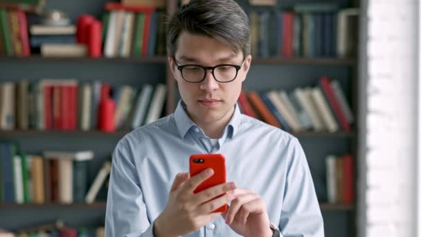 Молодой человек портрет с помощью смартфона смеется стоя в библиотеке социальные медиа смешно — стоковое видео