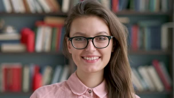 Portré, fiatal, csinos könyvtáros nő mosolyog, boldog látszó-on fényképezőgép-ban könyvtár könyvespolc háttér tudás tanulás közelről — Stock videók
