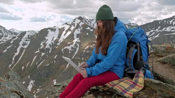 Mujer turista freelancer utiliza un ordenador en la naturaleza y se sienta en el césped junto a una antorcha de gas. Las montañas se reflejan en la pantalla del ordenador portátil — Vídeo de stock