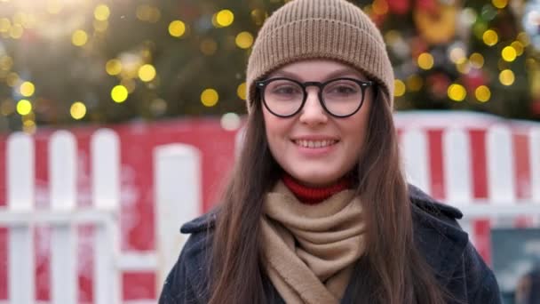 Mujer bonita se para en la plaza de la ciudad entre las decoraciones de Navidad y mira directamente a la cámara — Vídeo de stock
