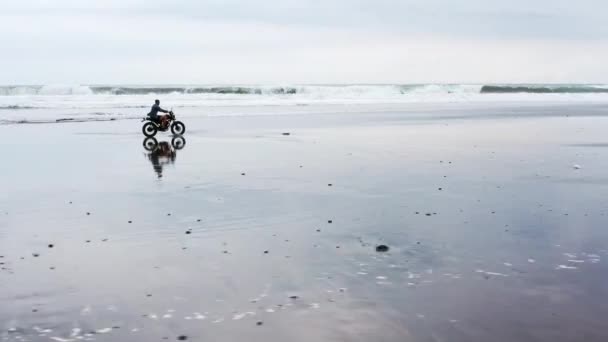 Jonge knappe hipster man paardrijden moderne custom motorfiets racer op het zwarte zandstrand in de buurt van het water. Surfplek met oceaangolven. — Stockvideo