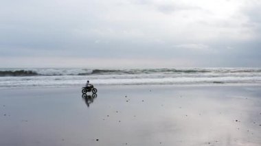 Genç yakışıklı hippi modern motosiklet yarışçısına suyun yanındaki siyah kumsalda biniyor. Okyanus dalgalı sörf noktası.