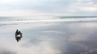 Genç yakışıklı hippi modern motosiklet yarışçısına suyun yanındaki siyah kumsalda biniyor. Okyanus dalgalı sörf noktası.