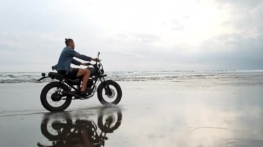 Genç yakışıklı hippi modern motosiklet yarışçısına suyun yanındaki siyah kumsalda biniyor. Okyanus dalgalı sörf noktası.