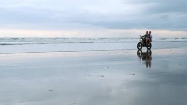 Genç, güzel bir çift hippi plajda retro motosiklet sürüyor, açık hava portresi, binici erkek ve kız, birlikte seyahat, okyanus, deniz