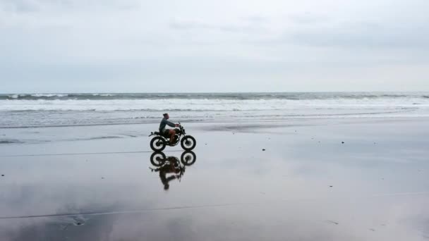 ビーチでオートバイに乗っている男。バリ島のビーチの夕日にヴィンテージバイク。自由とアクティブなライフスタイルを楽しむ若いヒップスターの男性は、バイカーツアーで楽しんでいます — ストック動画