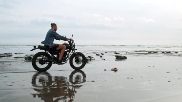 水の近くの黒い砂のビーチで現代のカスタムバイクレーサーに乗って若いハンサムなヒップスター男。海の波でサーフィンスポット. — ストック動画