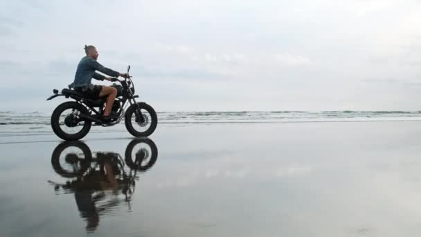 Ung stilig hipster man rider modern anpassad motorcykel racer på den svarta sandstranden nära vattnet. Surfingplats med havsvågor. — Stockvideo