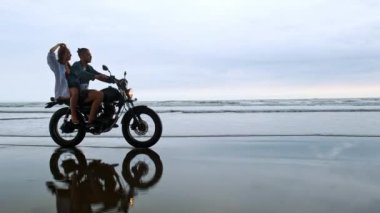 Genç, güzel bir çift hippi plajda retro motosiklet sürüyor, açık hava portresi, binici erkek ve kız, birlikte seyahat, okyanus, deniz
