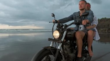 Genç, güzel bir çift hippi plajda retro motosiklet sürüyor, açık hava portresi, binici erkek ve kız, birlikte seyahat, okyanus, deniz
