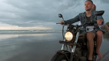 Genç, güzel bir çift hippi plajda retro motosiklet sürüyor, açık hava portresi, binici erkek ve kız, birlikte seyahat, okyanus, deniz