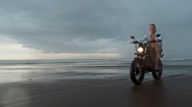 Sahilde motosiklet süren adam. Bali plaj günbatımı üzerinde vintage motosiklet. Genç hipster erkek özgürlük ve aktif yaşam tarzı zevk, bir bisikletçiler tur eğlenmek