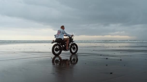 Genç yakışıklı hippi modern motosiklet yarışçısına suyun yanındaki siyah kumsalda biniyor. Okyanus dalgalı sörf noktası. — Stok video